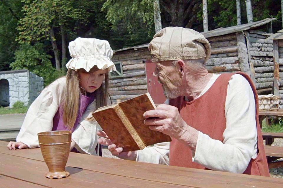 En bild som skildrar medeltiden, en man läser i en bok och flicka kikar fram över bordet. De har medeltida kläder och vistas i medeltida utomhusmiljö.