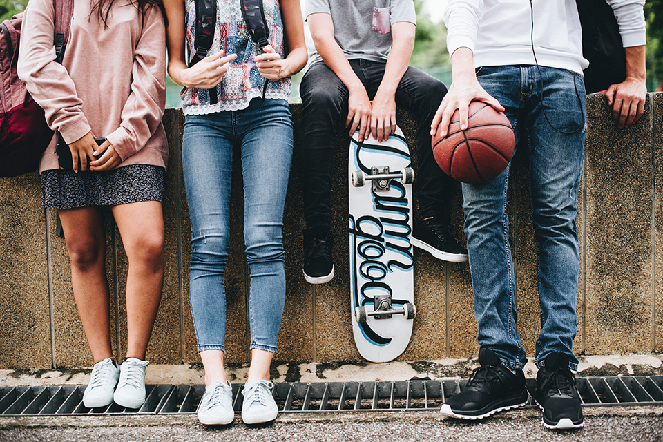 Benen på fyra ungdomar som sitter på en mur. De håller i mobil, basketboll och skateboard.
