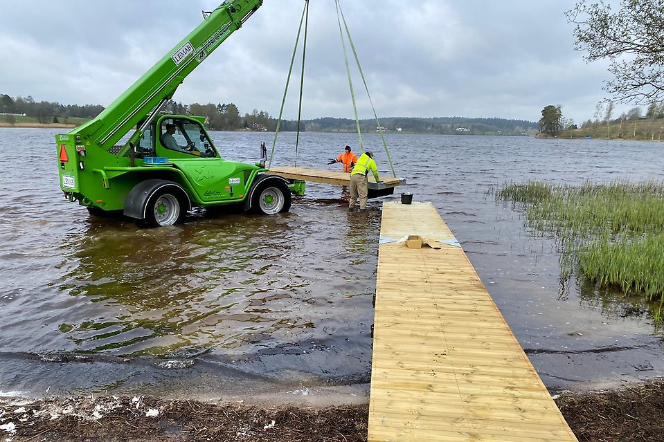 En grön bil med lyftkran lägger i en brygga i en sjö. Två personer står med vatten upp till vaderna och tar emot bryggan.