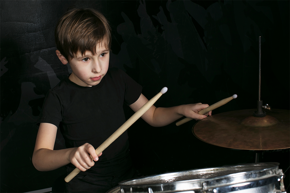 Pojke i mellanstadieåldern spelar trummor, rummet bakom är mörkt.