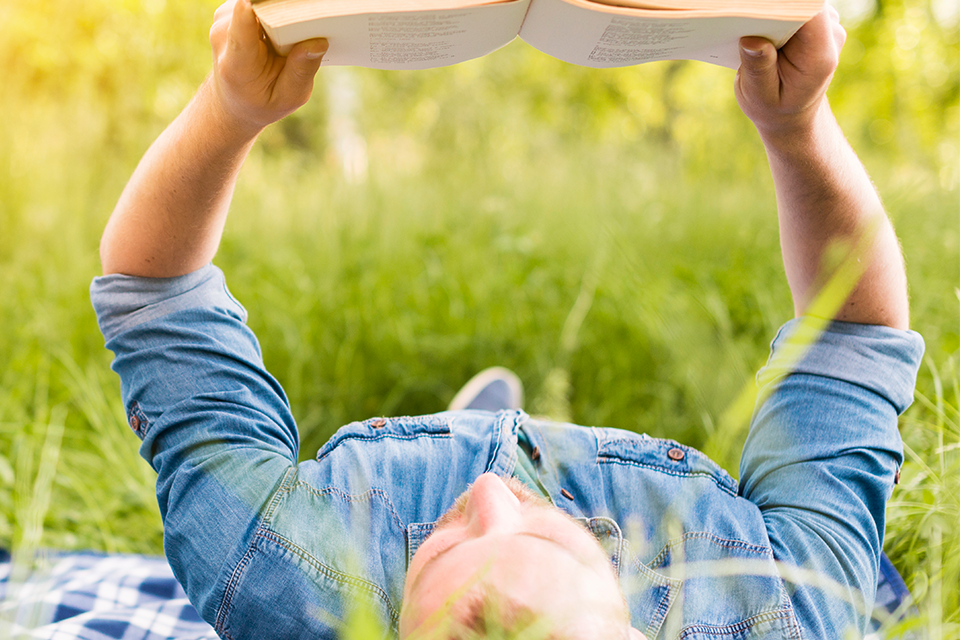 Man ligger i gröngräset på rygg och läser en bok.