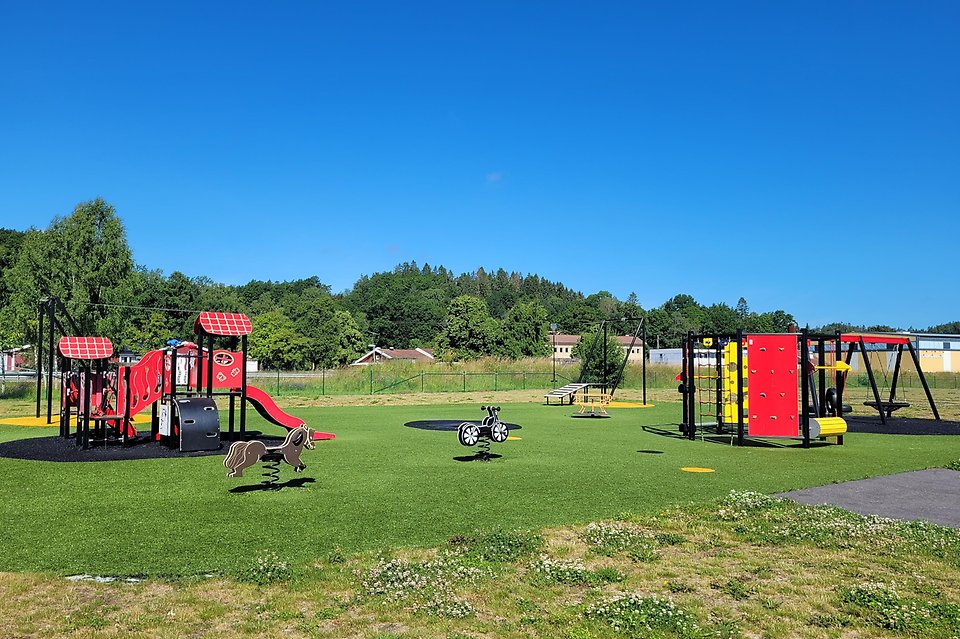 Bild från stor lekplats med underlag av grönt konstgräs. Det syns en klätterställning till vänster, en gungmotorcykel, en linbana, en stor rund aktivitetsställning till höger och en gunga i bakgrunden.