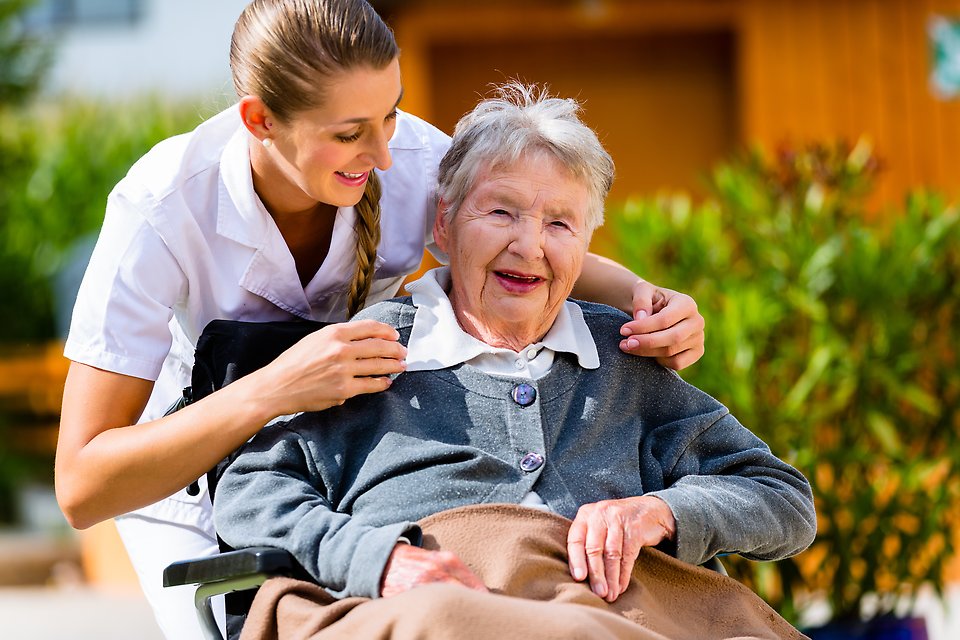 En kvinna i vit skjorta står bakom en äldre dam i rullstol