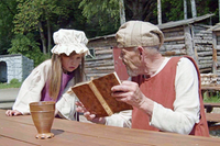 En bild som skildrar medeltiden, en man läser i en bok och flicka kikar fram över bordet. De har medeltida kläder och vistas i medeltida utomhusmiljö.