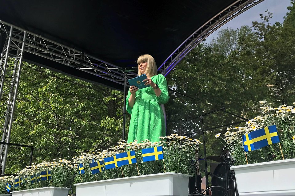 En blond kvinna i grön klänning håller ett tal på en scen. Framför henne syns blomkrukor med vita prästkragar och små vimplar med den svenska flaggan.