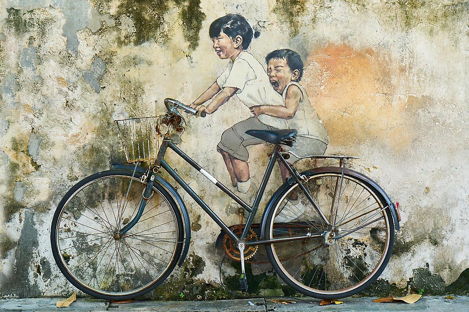 Gatukonst på mur, två barn cyklar, den ena skjutsar den andra på pakethållaren.