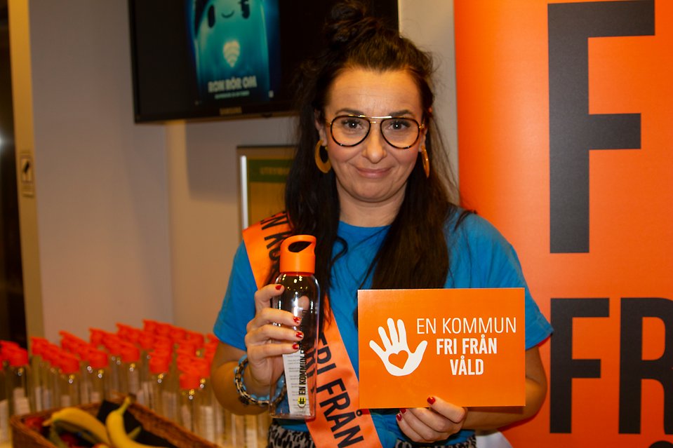 En kvinna håller upp en vattenflaska och ett orange vykort med texten "En kommun fri från våld"